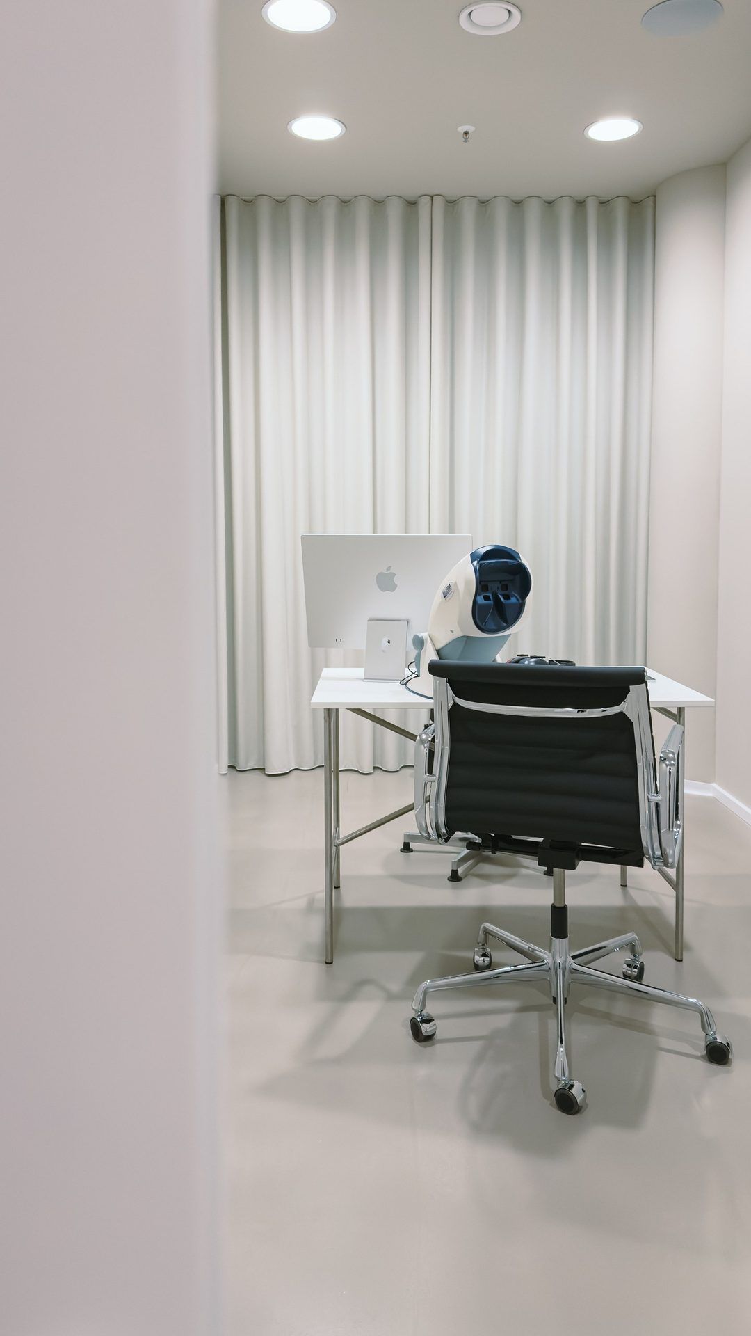 Bright examination room with a desk, a computer and a vision testing device, an office chair in front, curtains along the wall