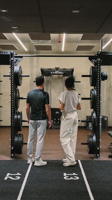Two people in front of a power rack with weight plates, lanes 12 and 13 marked on the floor