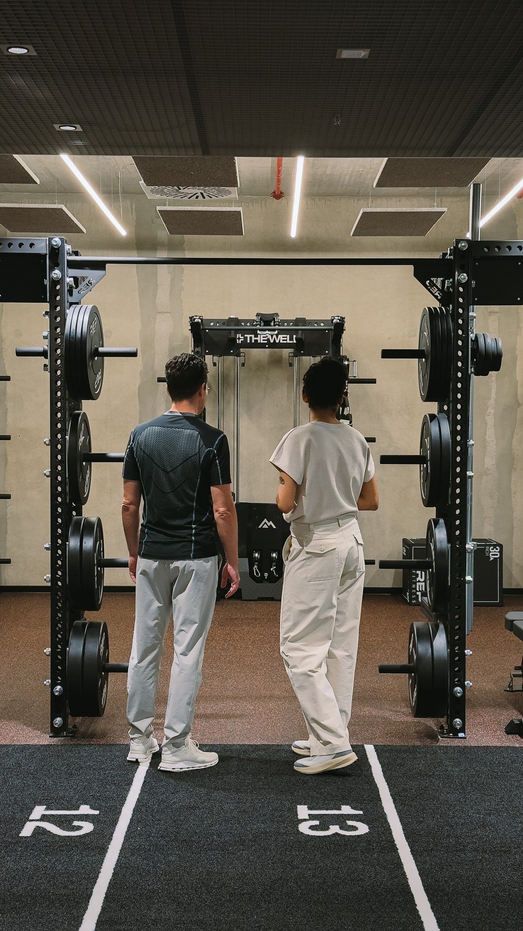Two people in front of a power rack with weight plates, lanes 12 and 13 marked on the floor