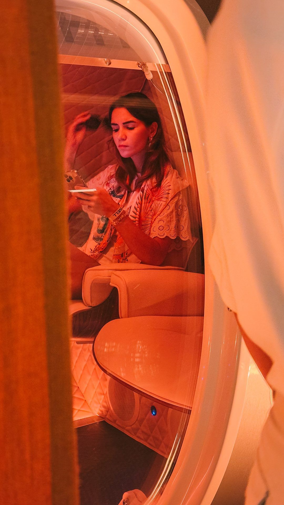 A woman sitting in the hyperbaric oxygen chamber, looking at her phone, seen through the open door in orange light