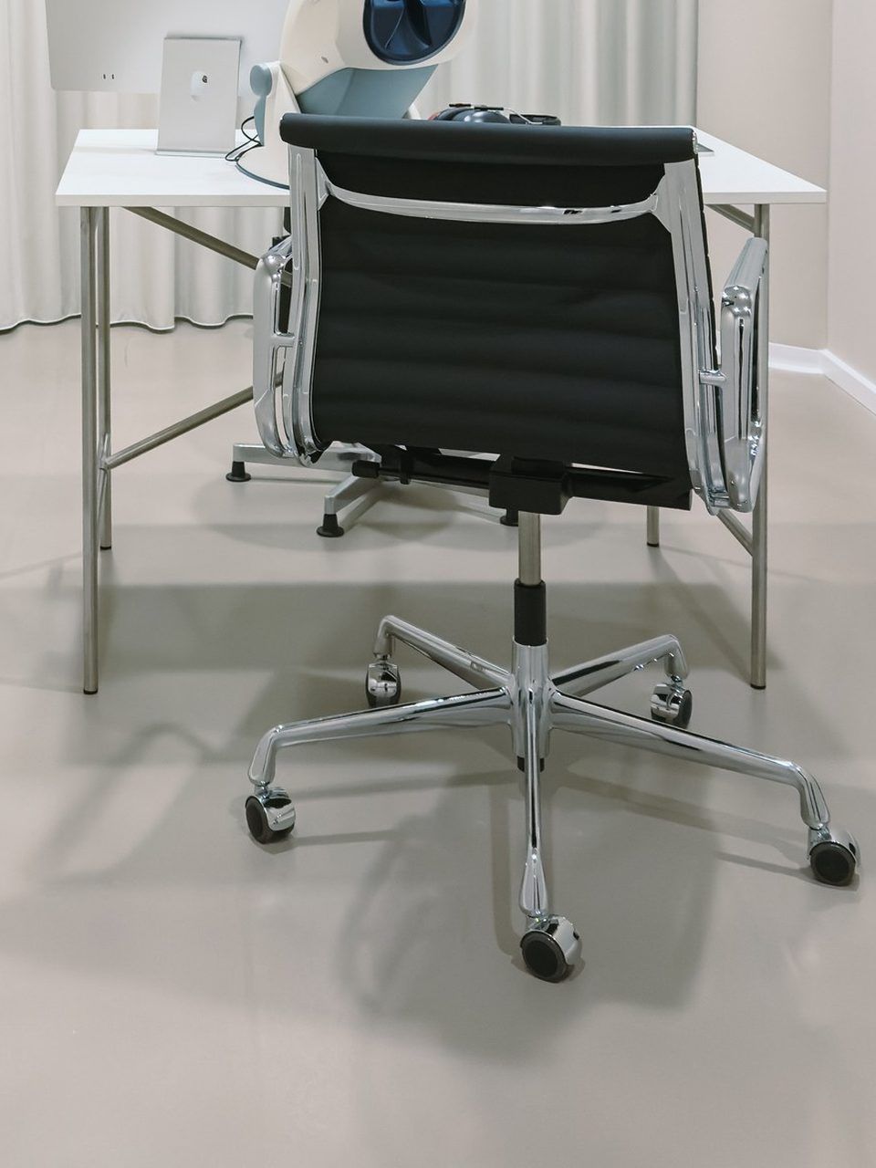 Office chair at a white desk in the examination room, a screen and the vision testing device on the desk, a light curtain behind