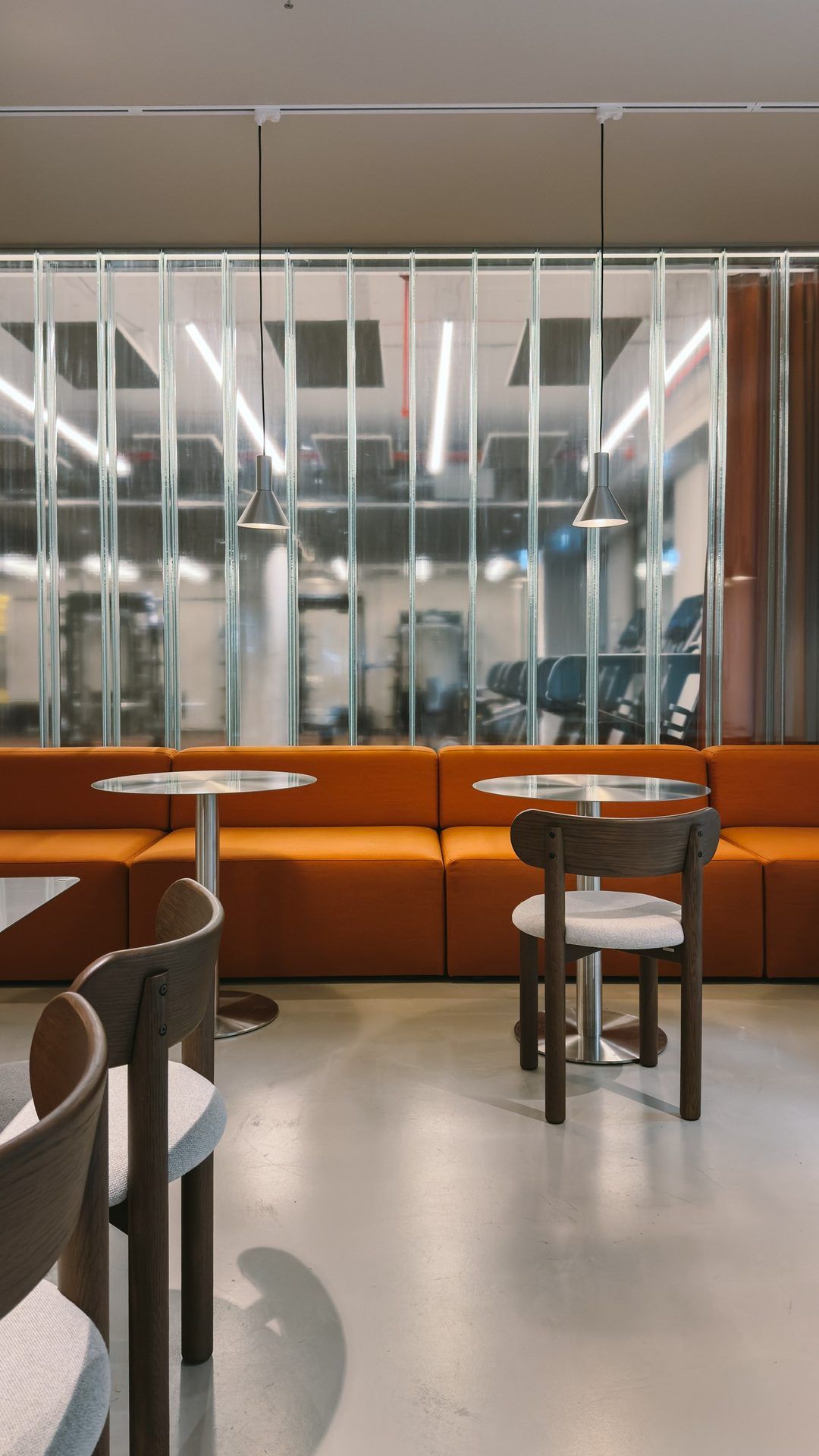 Lounge with an orange sofa and round bistro tables, the gym behind ribbed glass