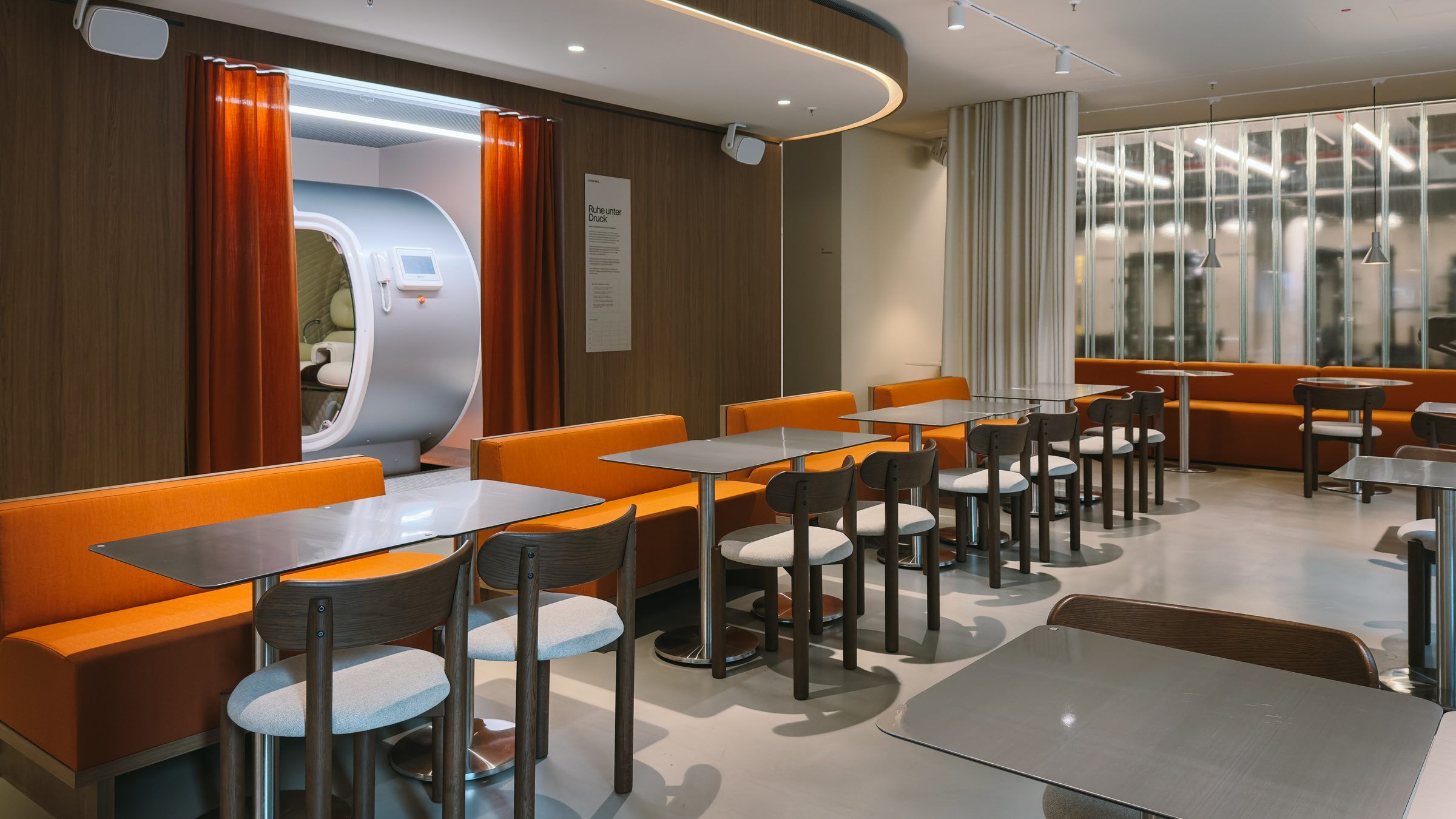 Restaurant with orange banquettes and bistro tables, the oxygen chamber alcove on the left and the gym behind fluted glass at the back