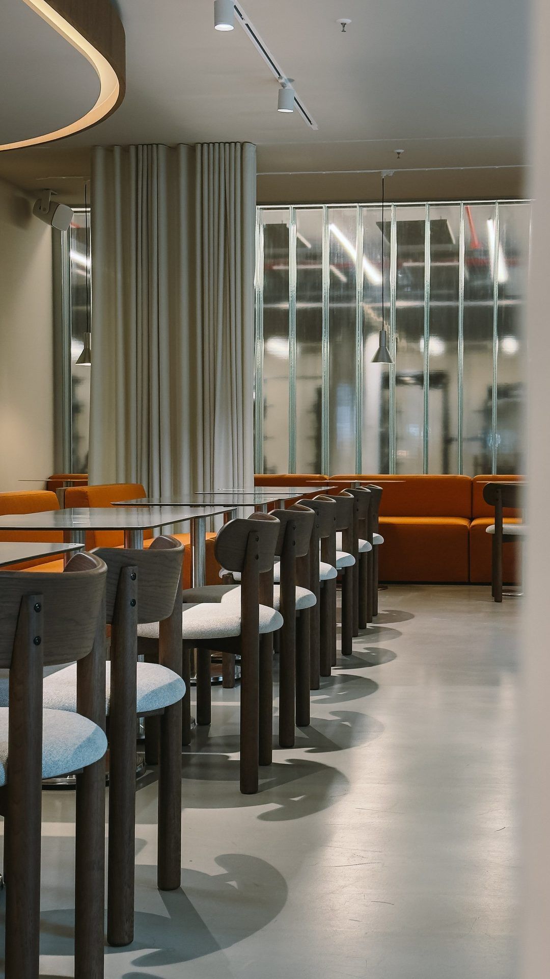 Restaurant with long rows of tables and wooden chairs, orange benches and a ribbed glass wall at the back