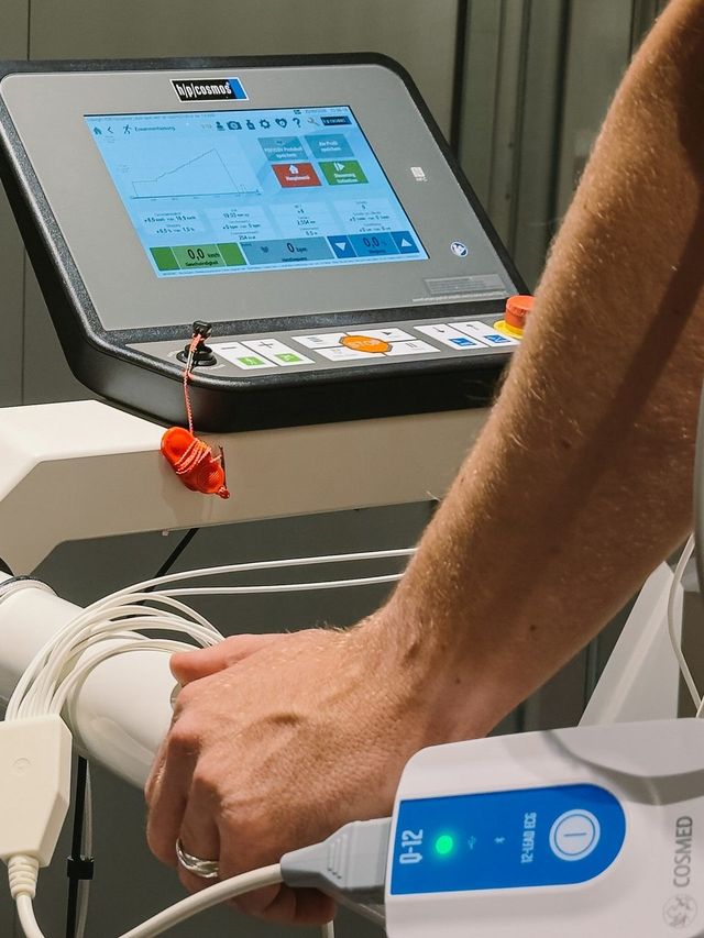 A hand on the handrail, below it the ECG device with its cables, above it the treadmill screen