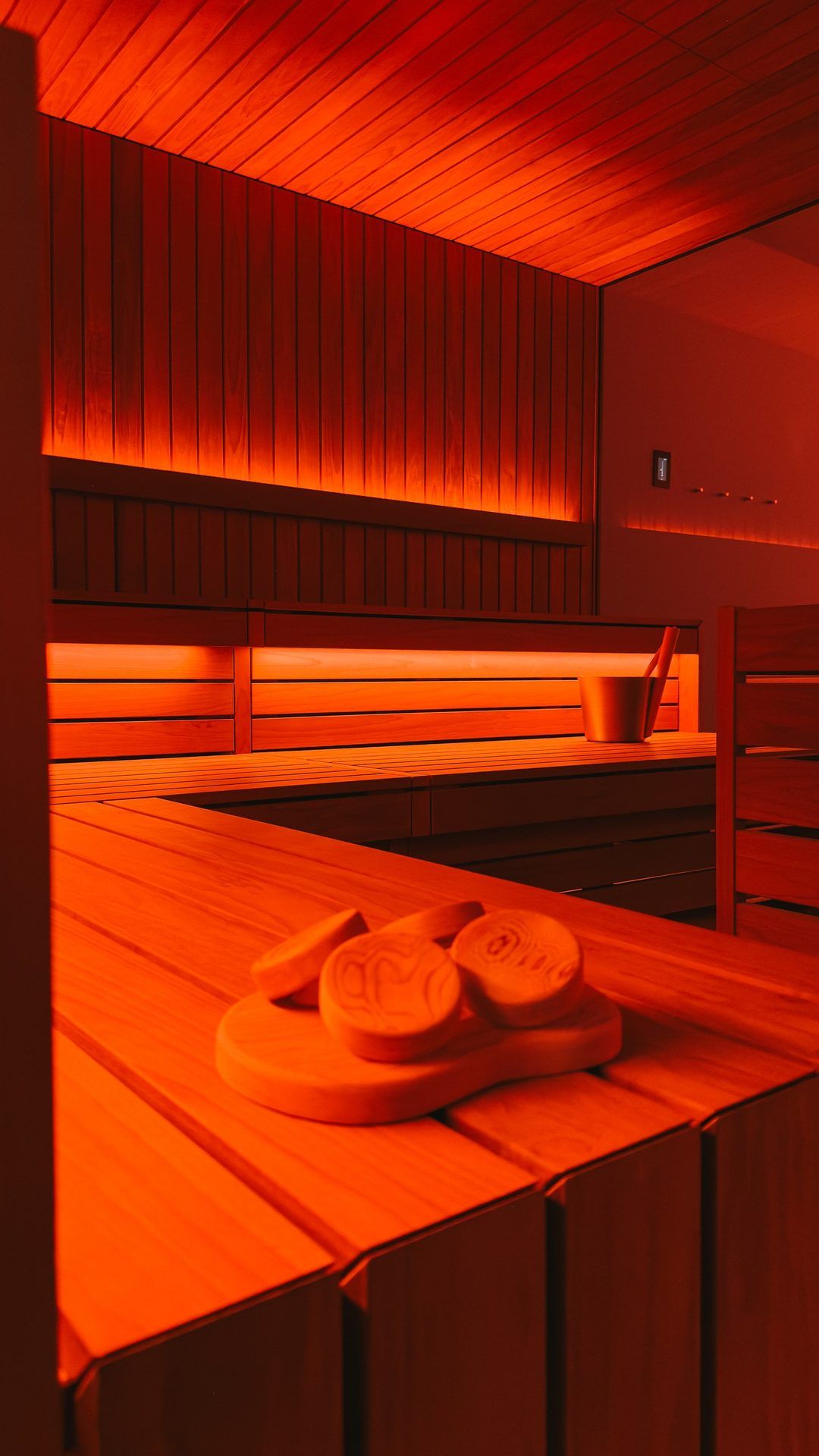 Sauna in red light with wooden benches, a pair of slides on the bench, a sauna bucket at the back